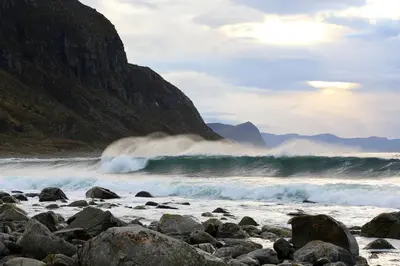 a wave crashing on a rocky beach next to a mountain at Alnes Gård in Ålesund
