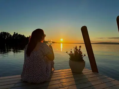 a woman sitting on a dock with a glass of wine watching the sunset at Idylliske Evjua ved Mjøsa in Skreia