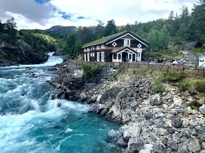a house on the side of a river with people standing outside at Billingen Seterpensjonat in Skjåk